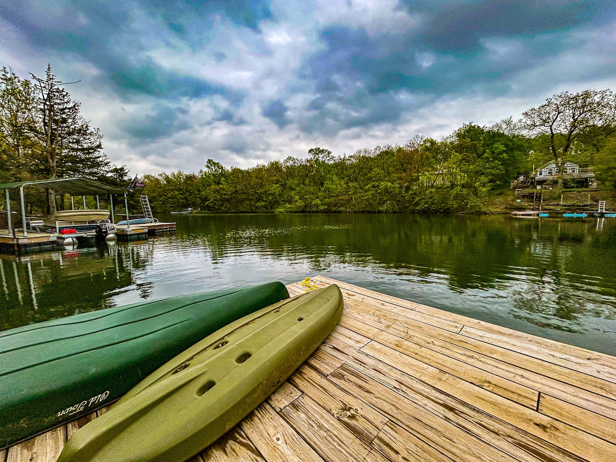 Two green kayaks rest on a wooden dock, with calm water extending beyond. Lush greenery lines the shoreline, with trees reflecting on the surface. A cloudy sky adds a touch of serenity to the lakefront scene.