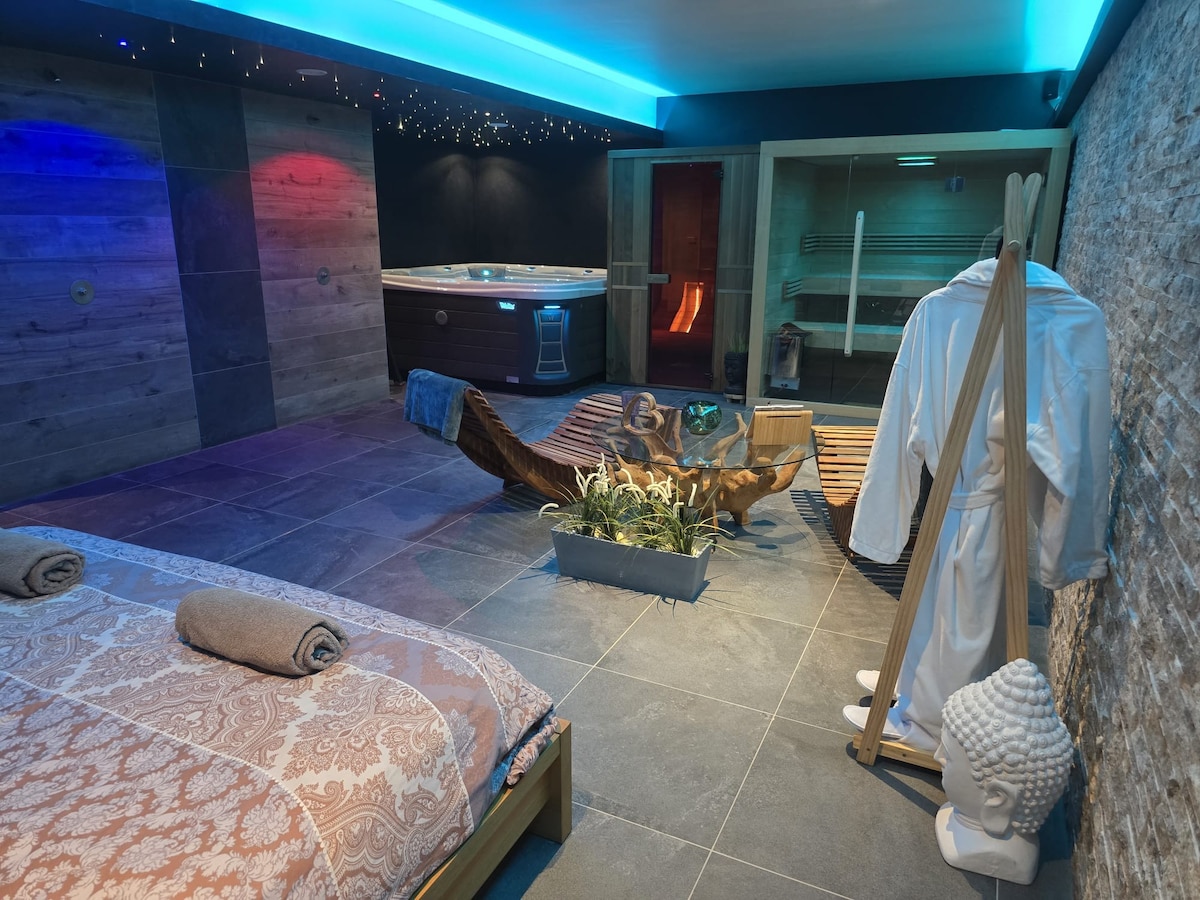 An inviting wellness area features a hot tub, sauna, and infrared cabin, all surrounded by stone walls and soft lighting. A comfortable lounge chair and a cozy bed with elegant linens are present. Decorative elements include a plant and a statue, enhancing the serene environment.
