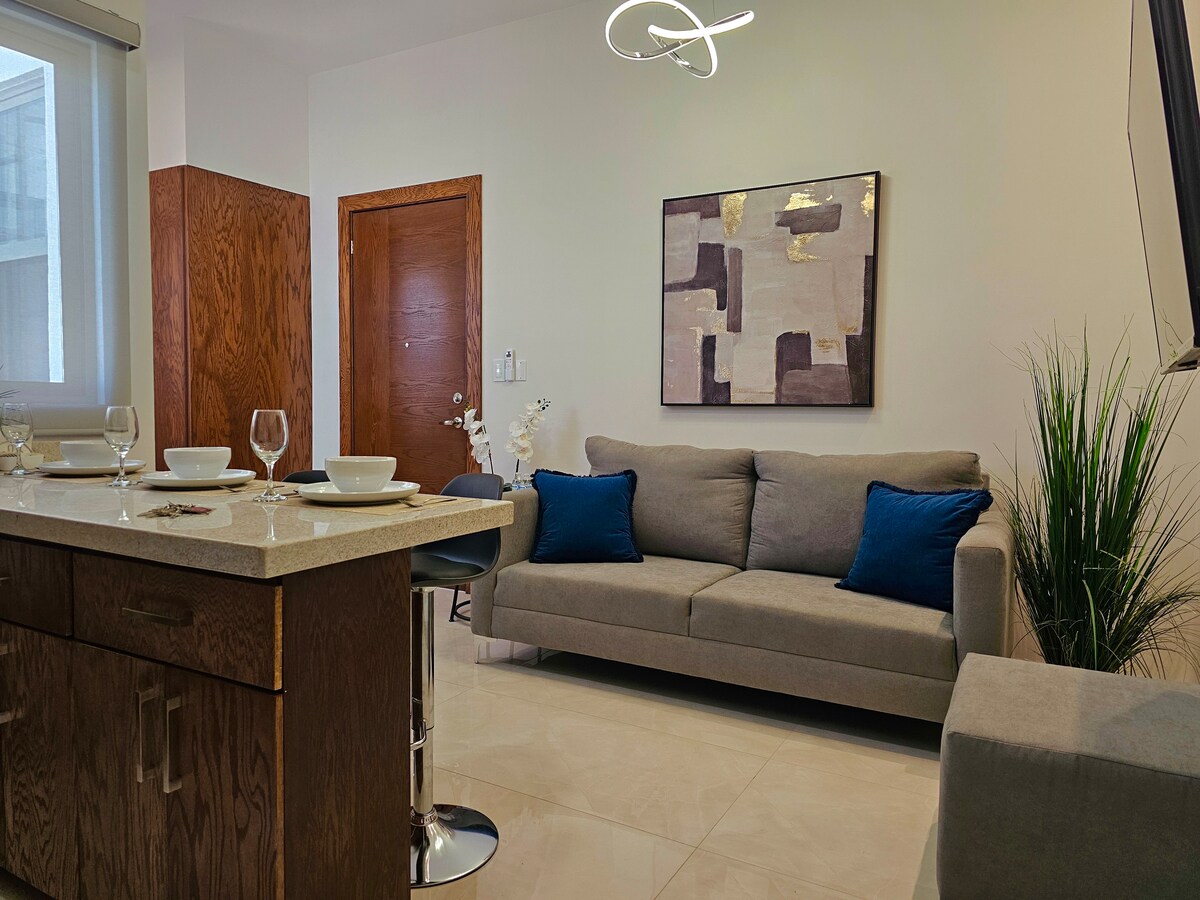 A modern living area features a light-colored sofa with two blue accent pillows, complemented by a sleek coffee table. A stylish abstract wall art piece hangs above the sofa. A well-equipped kitchen island with dining settings is visible, enhancing the open concept layout.