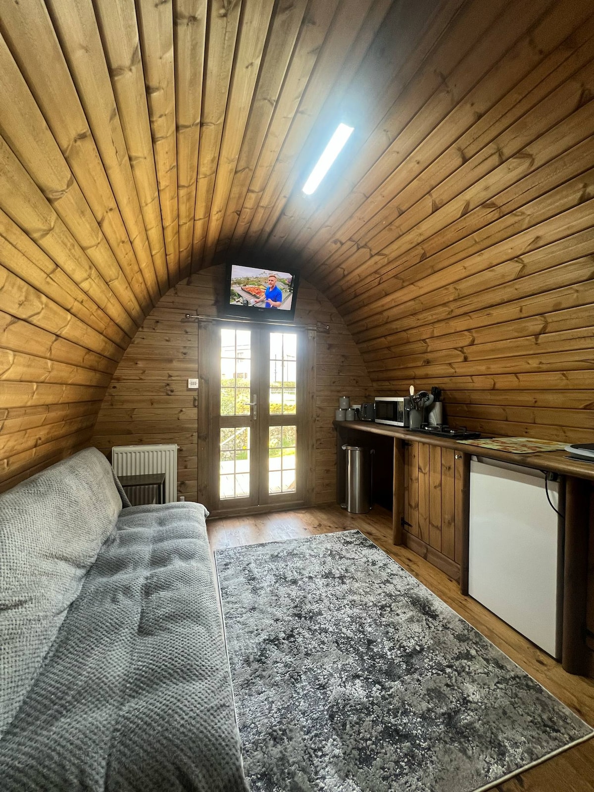 A cozy interior features a curved wooden wall, providing a rustic aesthetic. A gray sofa is positioned in front of a kitchenette, which includes a microwave and refrigerator. A small television is mounted on the wall, while a door leads to the outdoor area, allowing natural light to filter in.