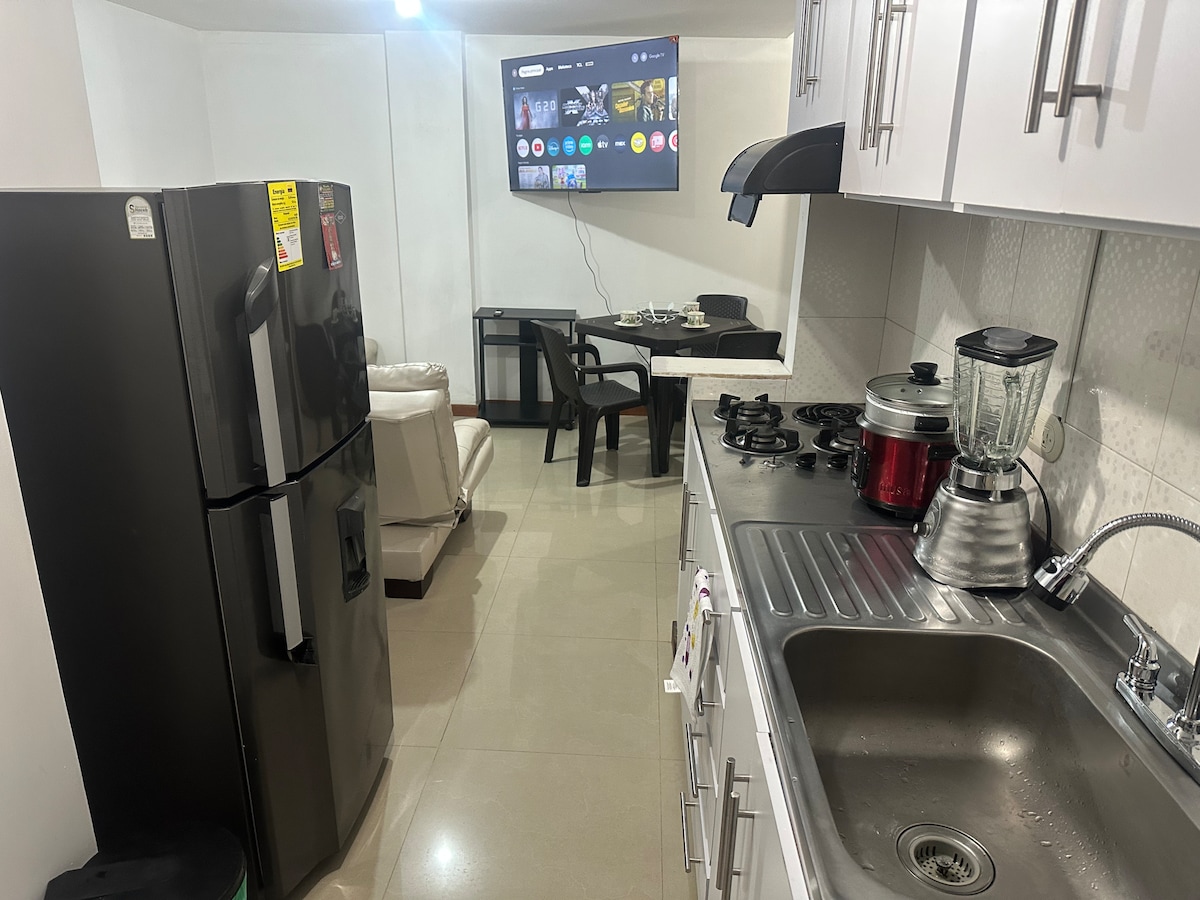 A functional kitchen area features modern appliances, including a large refrigerator and a stovetop with multiple burners. A wall-mounted television is visible in the space, accompanied by a small dining table and a cozy seating arrangement with a light-colored couch.