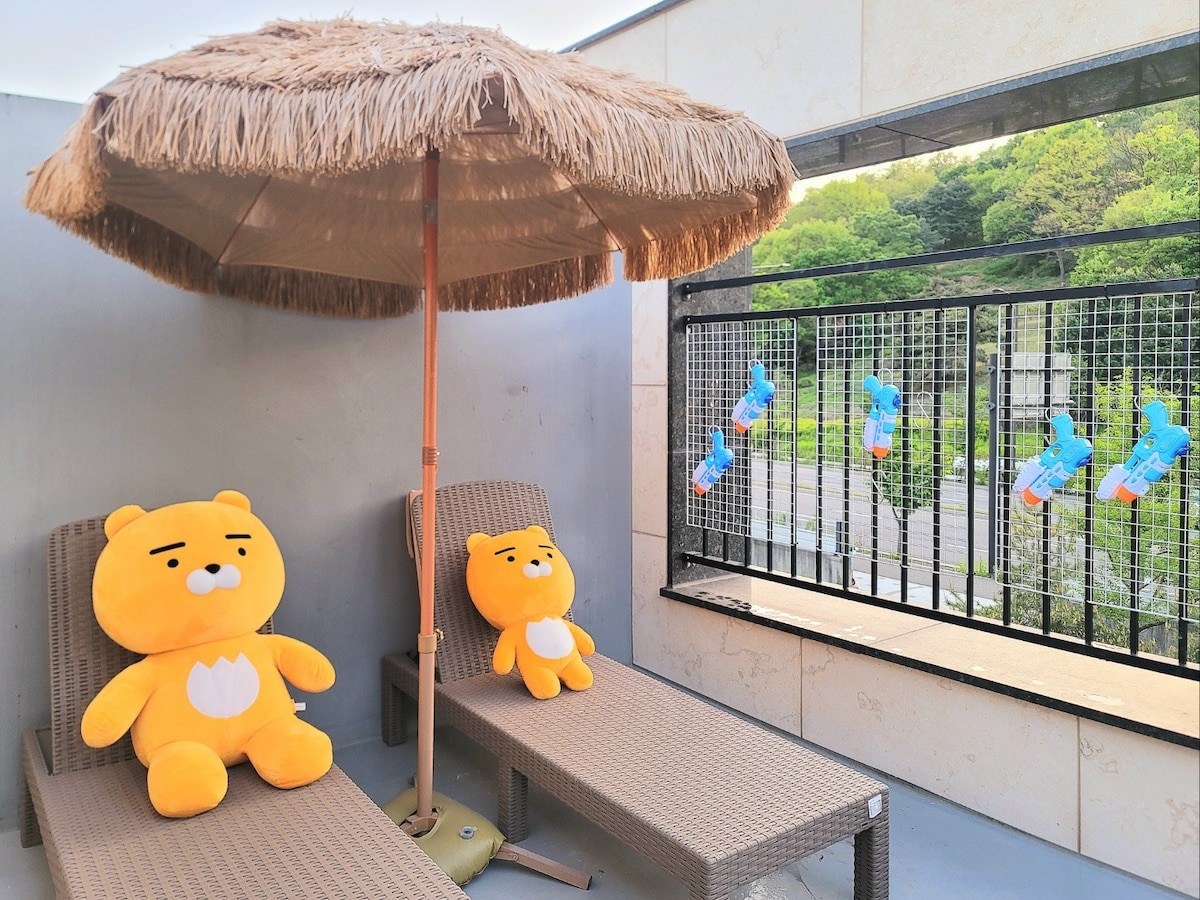A cozy outdoor terrace features two comfortable lounge chairs beneath a thatched umbrella. Plush yellow bear figures are positioned on the chairs. A playful arrangement of water guns hangs from the railing, adding a fun element to the serene natural setting in the background.