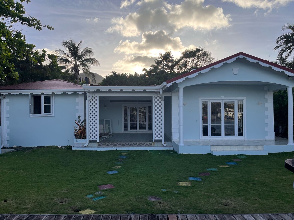 A charming exterior view features a light blue house with a red roof. The spacious front yard is adorned with green grass and a pathway lined with colorful stepping stones. Large glass windows allow natural light to flood the interior spaces.