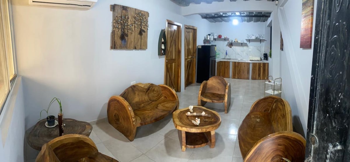 A cozy living space is furnished with unique, handcrafted wooden seating and a round coffee table. Light-colored walls and tiled flooring enhance the airy feel, while the kitchen area is visible in the background, featuring modern appliances and wooden cabinetry.