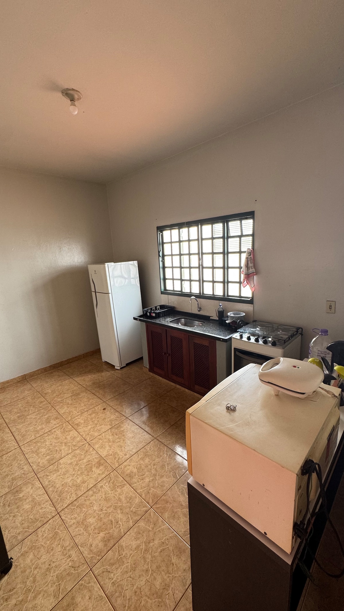 The kitchen space features light-colored walls and a tiled floor. A refrigerator and a gas stove are positioned along one wall, while a window allows natural light to enter. Counter space is visible, providing ample room for preparation and cooking.