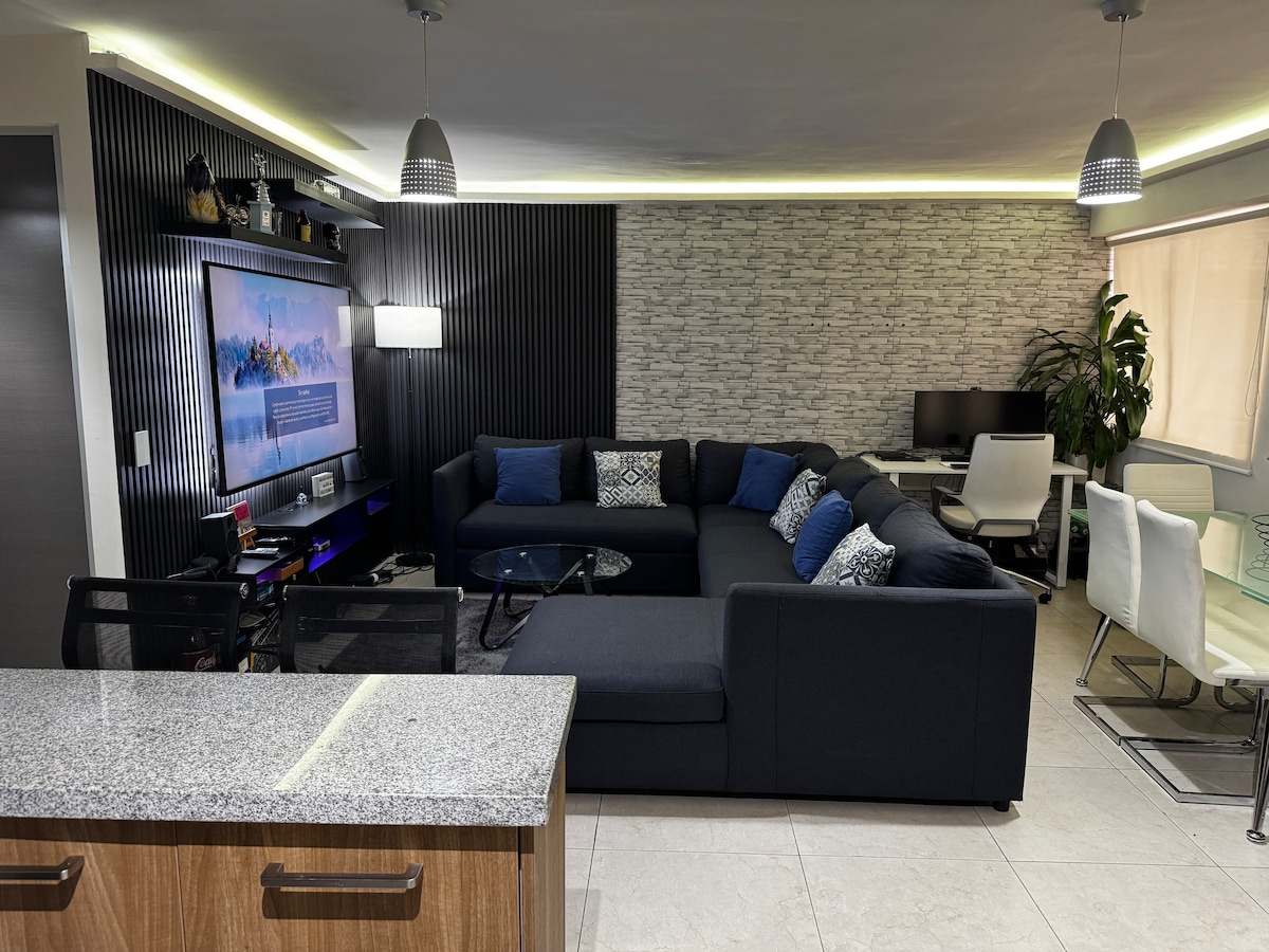 A modern living area features a sectional sofa adorned with blue and gray cushions. A large TV is mounted on the wall opposite, complemented by soft lighting and a textured backdrop. A glass-top table sits in front of the sofa, inviting relaxation.