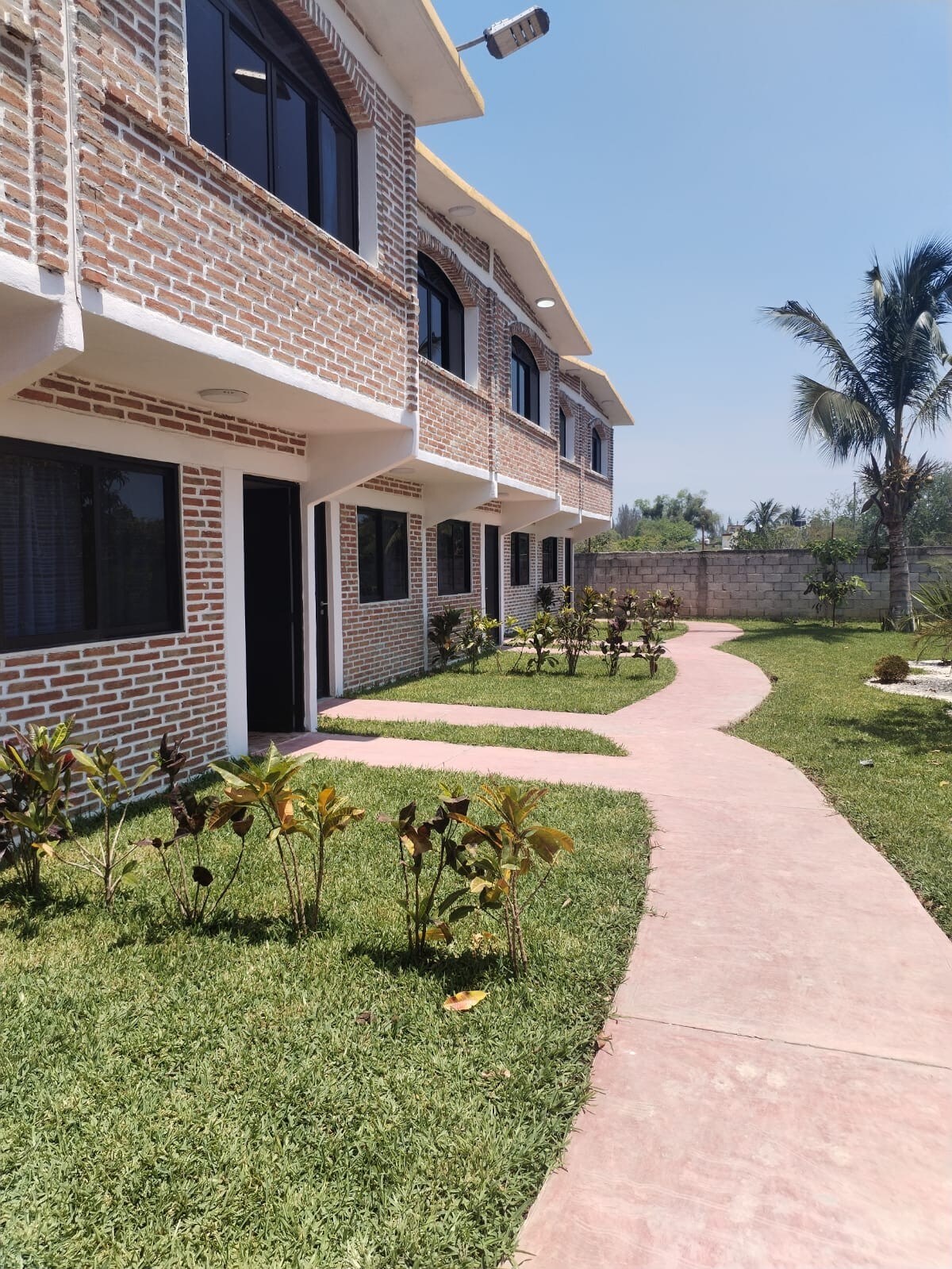 A well-maintained pathway winds through lush green grass, leading to a series of bungalows with textured brick exteriors. Large windows allow natural light to fill the spaces, while palm trees stand nearby, enhancing the serene outdoor environment.