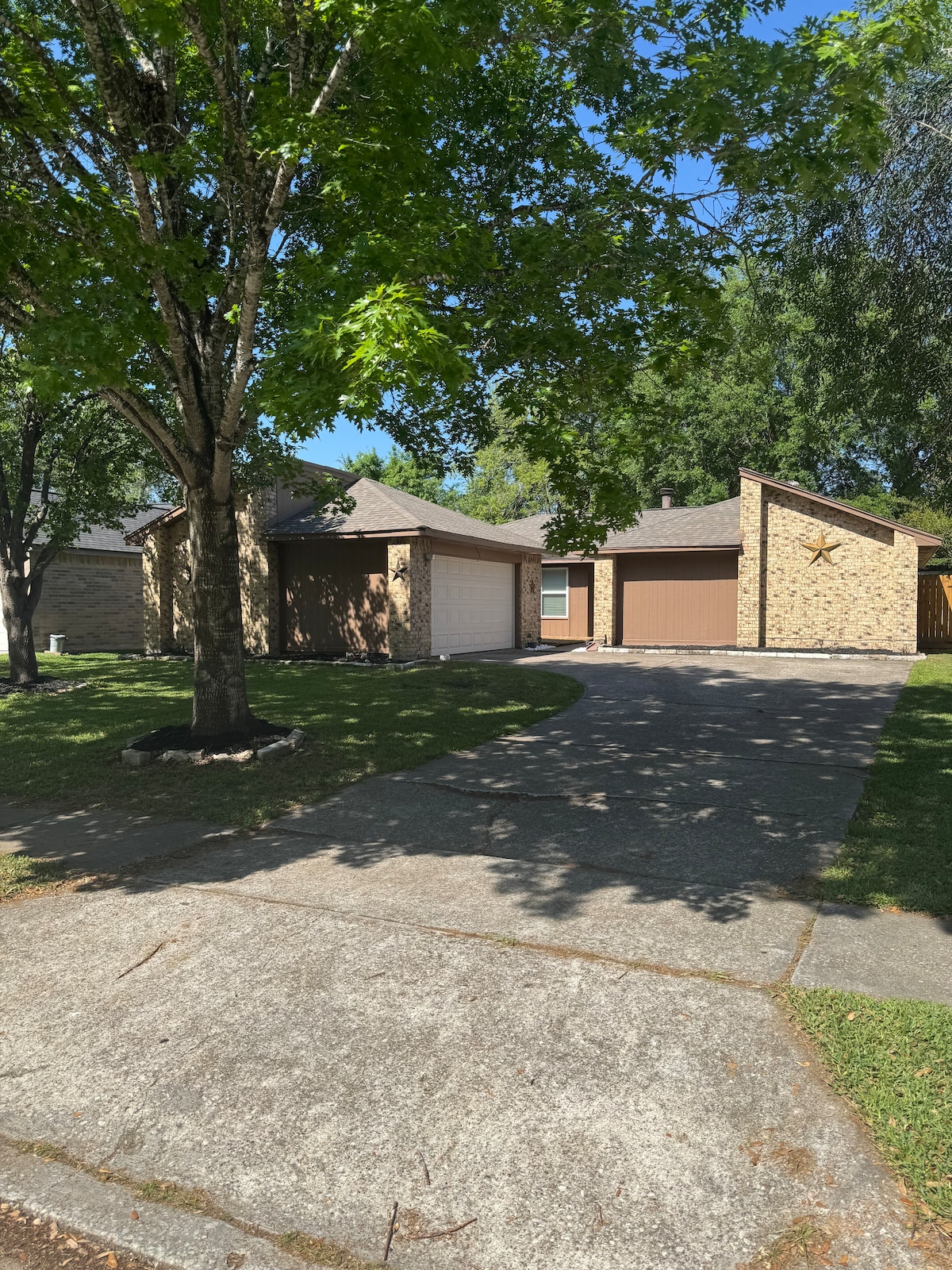 The exterior of a well-maintained single-story home is displayed, featuring a mix of stone and brick materials. A two-car driveway leads to an attached garage. Tall trees provide shade over the front yard, with a manicured lawn contributing to the inviting atmosphere.