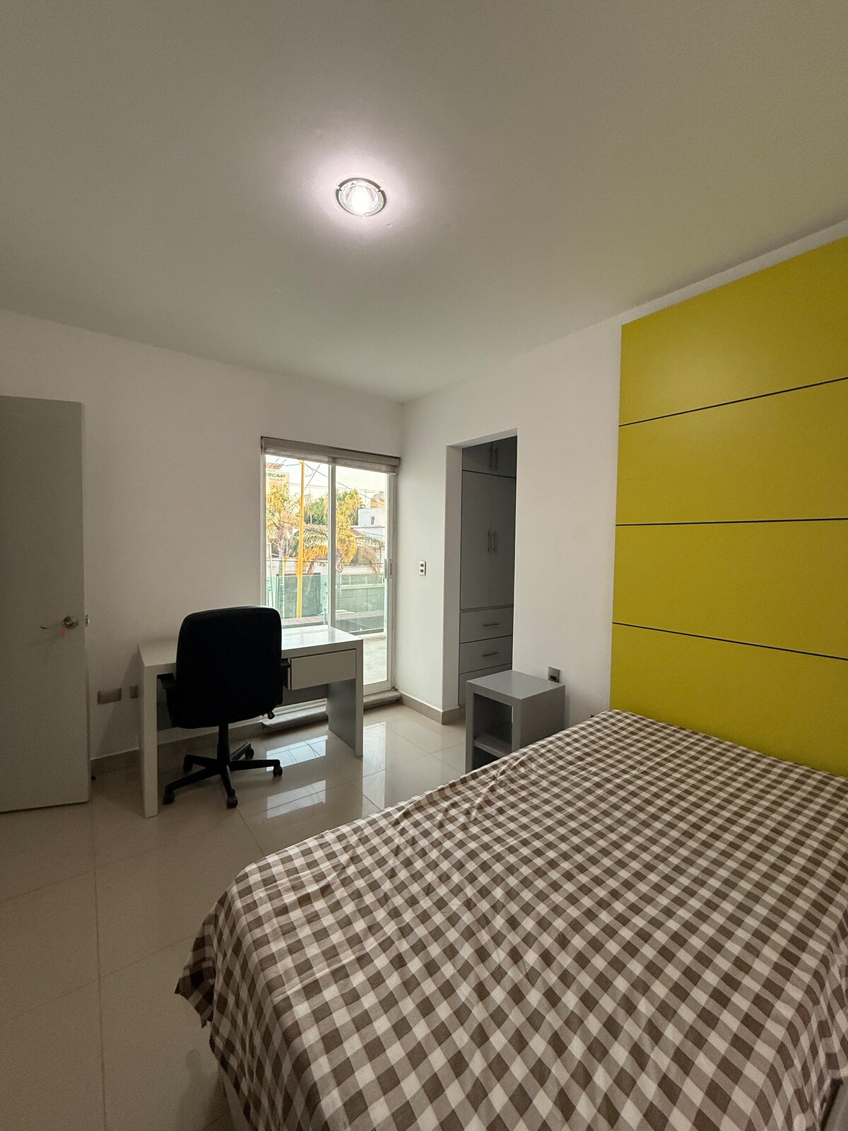 A well-lit bedroom features a spacious bed adorned with a checkered blanket. A desk and chair are positioned near a window, allowing natural light to enter. A yellow accent wall adds a pop of color, while light tiles enhance the room's brightness.