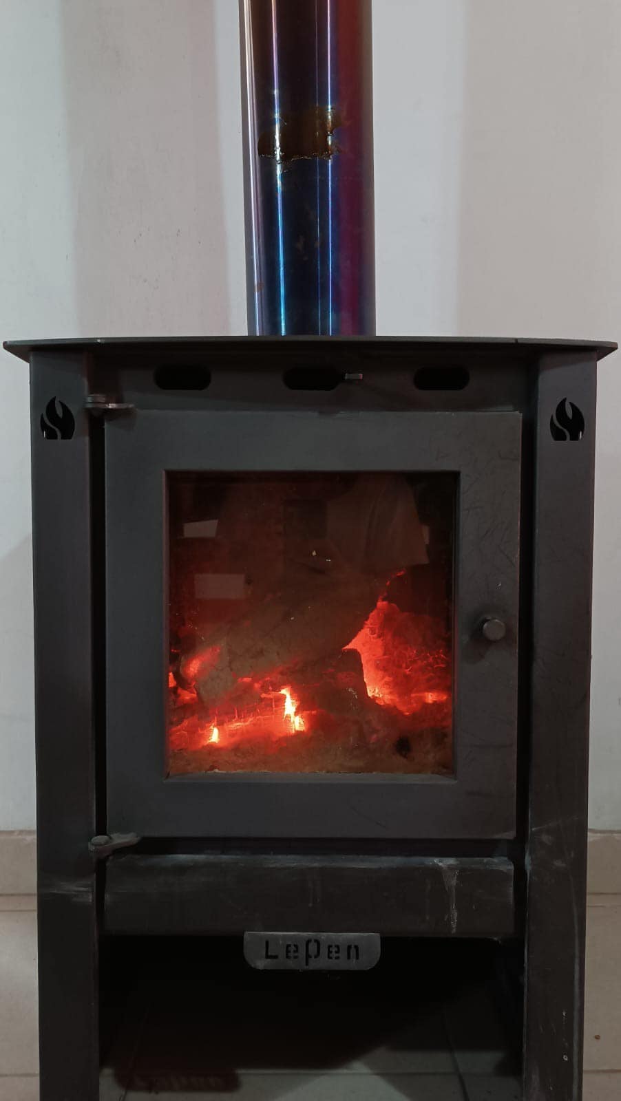 A wood-burning stove is positioned against a white wall, featuring a glass front showing glowing embers inside. A metallic chimney extends upward, presenting a contrasting effect with the stove's dark exterior. The tiled floor complements the overall cozy ambiance of the space.