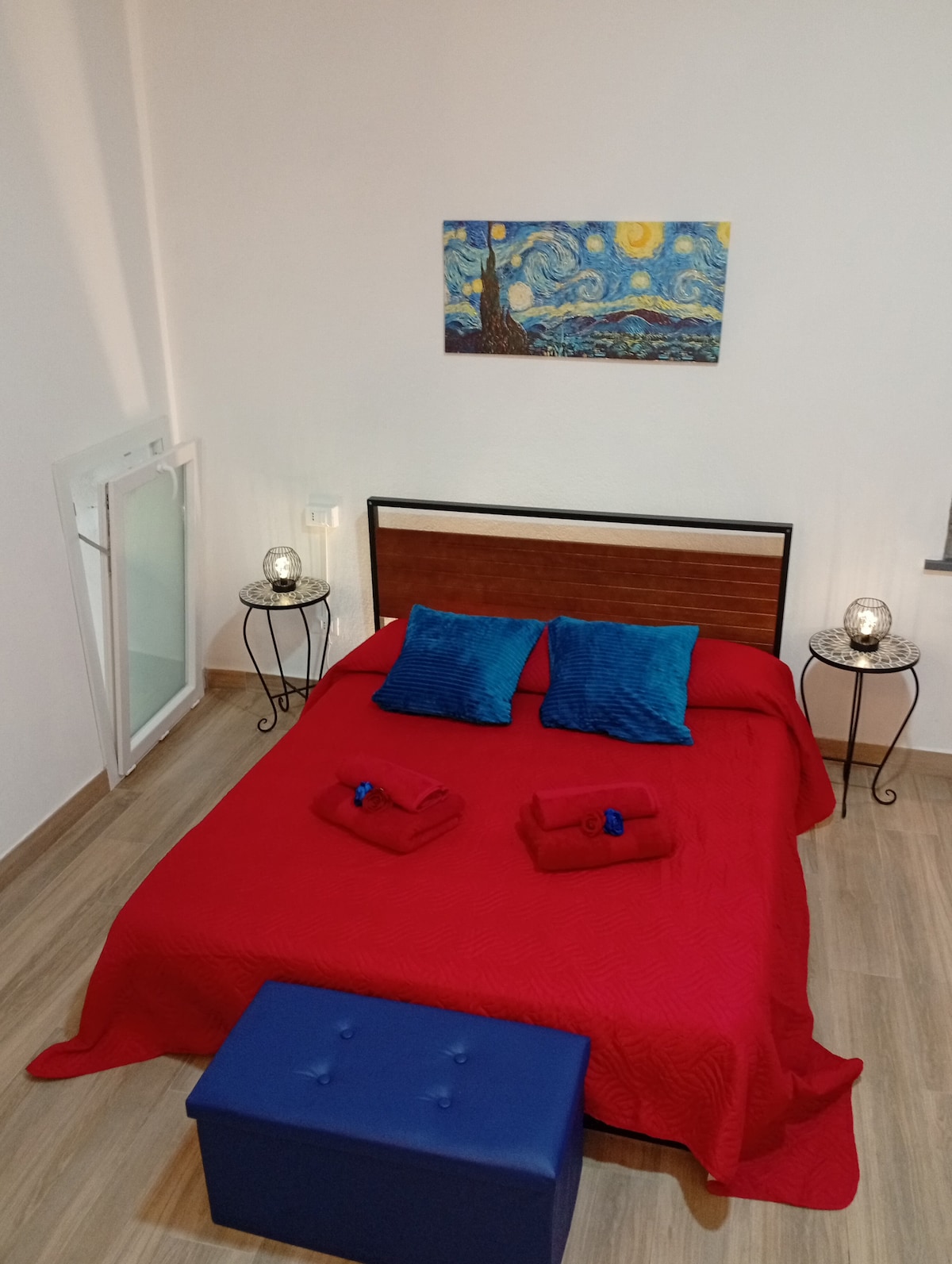 A spacious sleeping area is arranged with a vibrant red bedspread featuring pillows in blue hues. A simple blue storage bench sits at the foot of the bed. Two side tables with decorative lanterns enhance the setting, complemented by a piece of artwork on the wall.