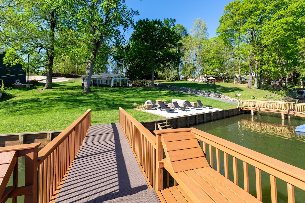 A wooden dock extends over calm waters, leading to a grassy area with a gently sloping yard. Lounge chairs are positioned on a sandy beach area nearby, surrounded by lush trees that provide shade. A sunlit space invites relaxation by the lake.