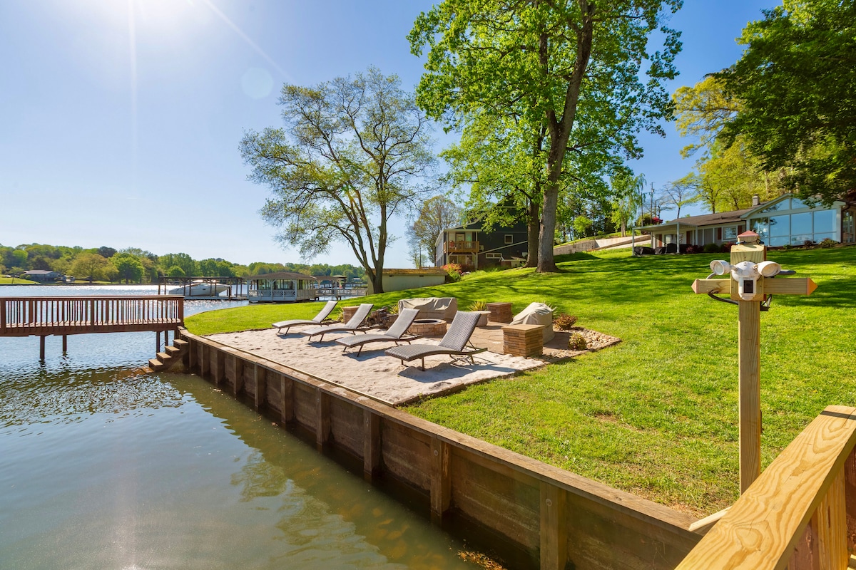 A serene lakeside area is presented, featuring a sandy beach with lounge chairs for relaxation. A grassy yard extends up to a private dock, while the clear water reflects the sunlight. Mature trees provide shade, enhancing the natural ambiance of the setting.