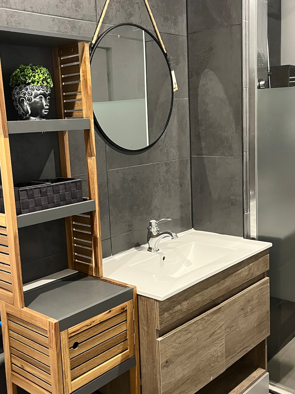 A modern bathroom space is displayed, featuring a sleek vanity with a white sink and storage drawers. A circular mirror hangs above the sink, and a wooden shelving unit with decorative elements is positioned beside it. A glass shower stall is visible in the background.