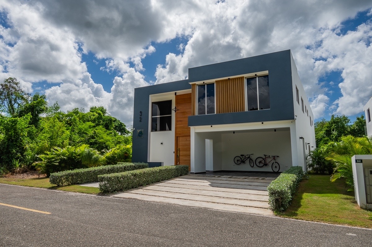 A modern two-story villa is set against a clear blue sky with fluffy clouds. Lush greenery frames the property, enhancing the curb appeal. A paved driveway leads to the entrance, with bicycles parked under the covered area. The exterior features a clean design with large windows.