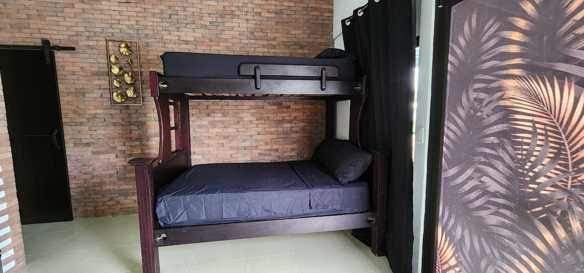 A bunk bed is featured in a room with a brick accent wall. The lower bunk is dressed in dark bedding, while the upper bunk rests above, both framed by dark curtains. A nearby decorative panel displays a tropical design.