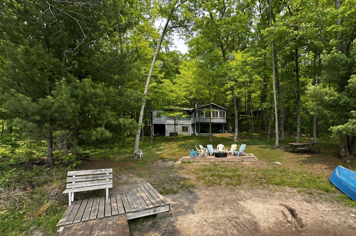 The outdoor space features a clearing surrounded by trees, with a cozy cabin visible in the background. Several lounge chairs are arranged around a firepit, and a wooden dock leads into a sandy area. A picnic table sits nearby, ideal for family gatherings.