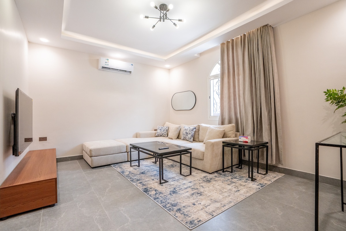 The living room displays a spacious, light-toned sectional sofa paired with a modern coffee table on a textured rug. A sleek wall-mounted TV is positioned opposite the seating area. Natural light enters through the arched window, complemented by soft drapes.