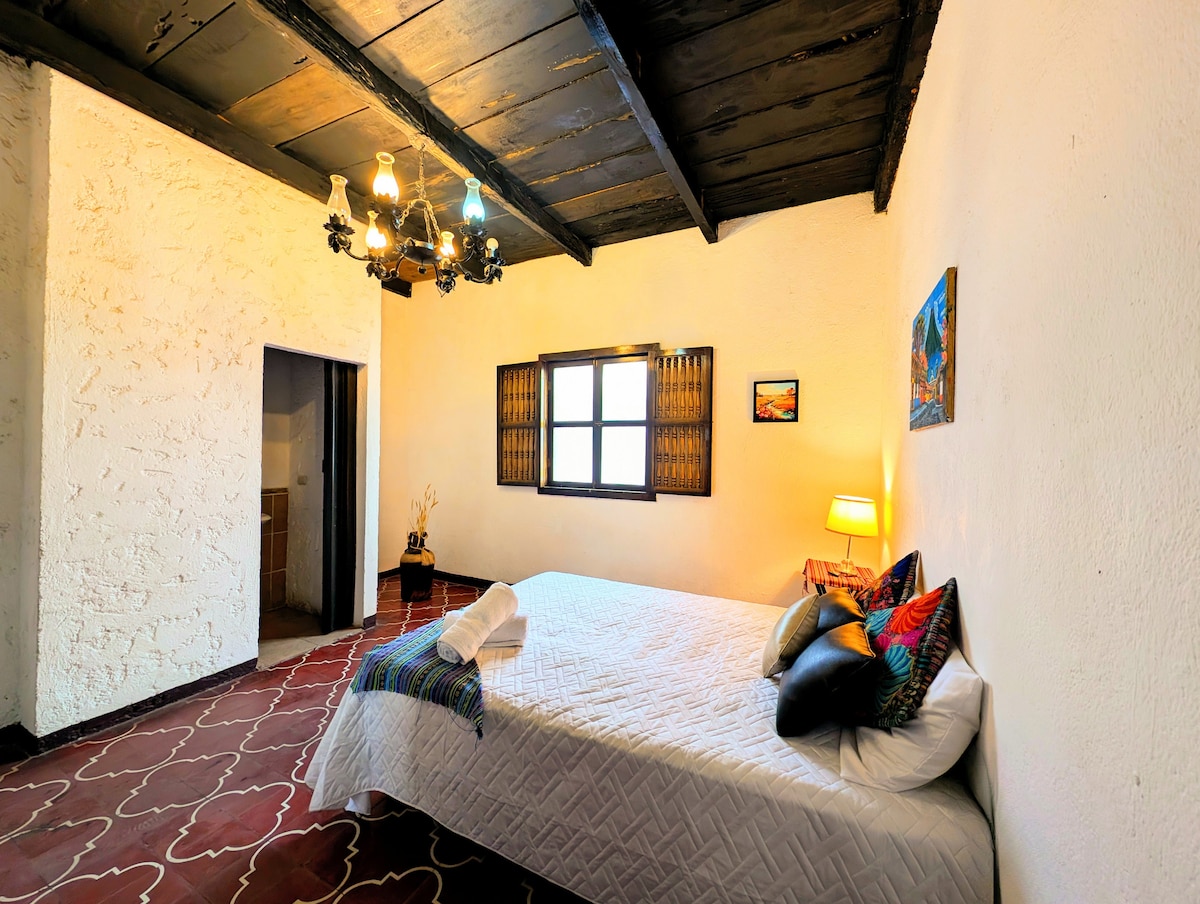 A spacious room features a large bed adorned with decorative pillows. A soft, white blanket is neatly arranged over the bed. An artistic wall hanging and a bedside lamp add character, while a wooden ceiling contrasts with the light-colored walls. A window with wooden shutters allows natural light to enter.