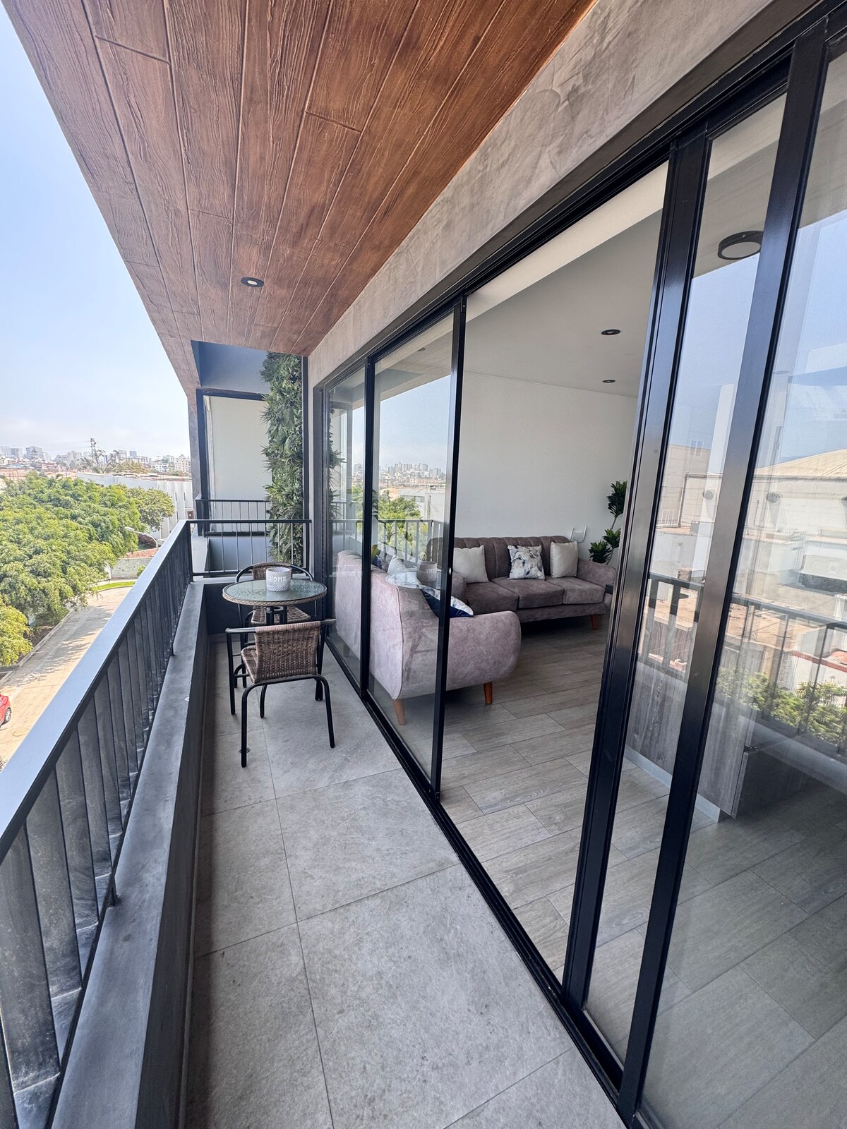 A spacious balcony is shown, featuring a glass railing that offers views of the surrounding greenery. Two comfortable chairs and a small table are positioned on the balcony, inviting relaxation. Natural light fills the area, enhancing the overall ambiance with a bright and airy feel.