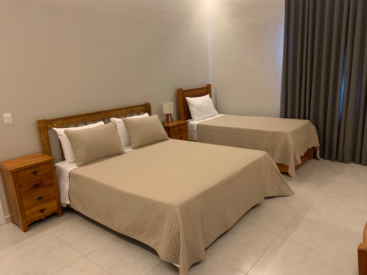 A spacious guest room is furnished with a queen size bed and a single bed, both draped in neutral-toned blankets. Wood nightstands are positioned beside each bed, and sunlight is softly filtered through a large window dressed with dark curtains.