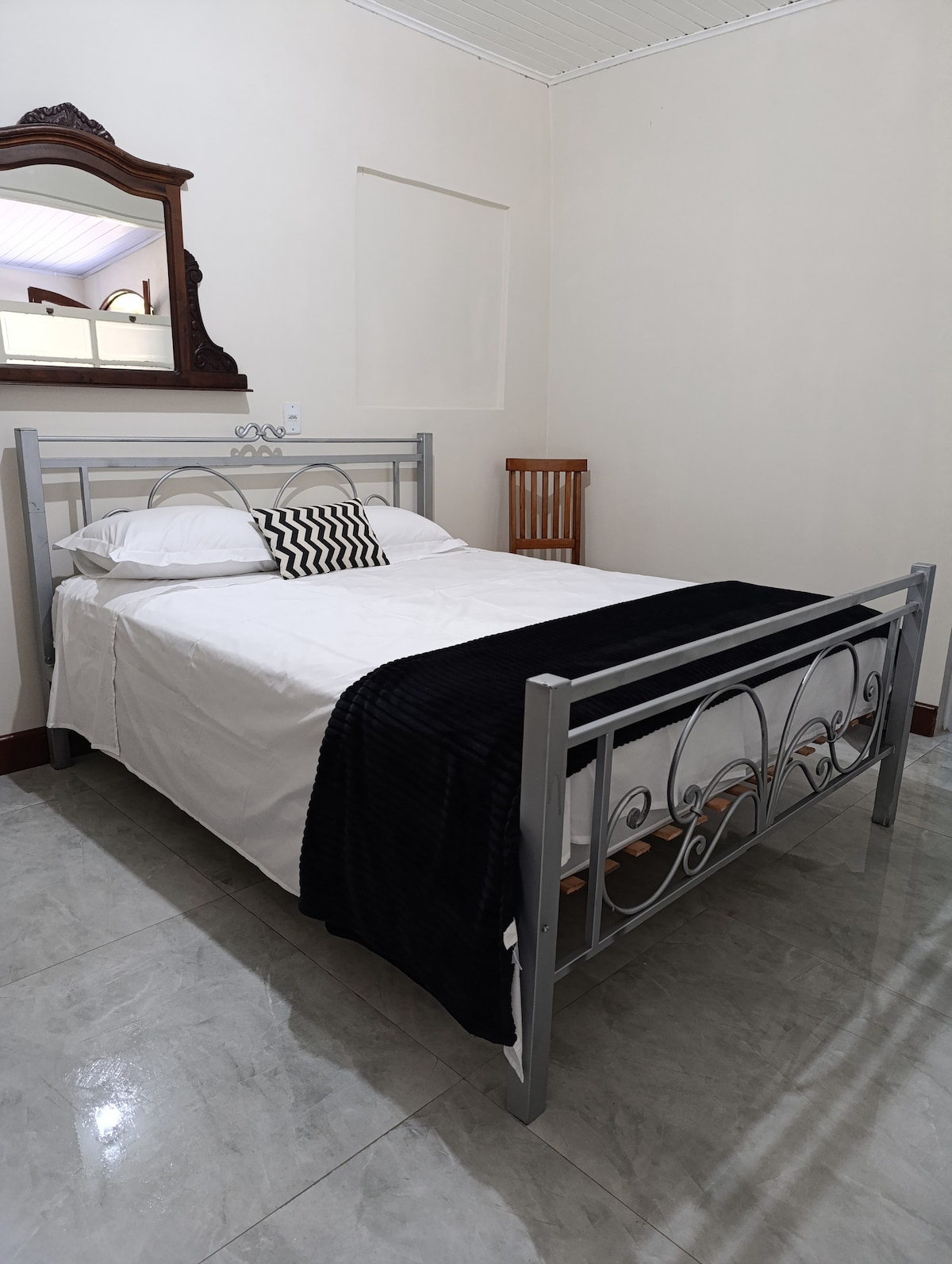A sleeping area features a metal framed bed with a white bedspread and a black throw blanket. A striped cushion adorns the bed. A wooden chair and a mirror are positioned nearby, with light reflecting softly off the tiled floor.