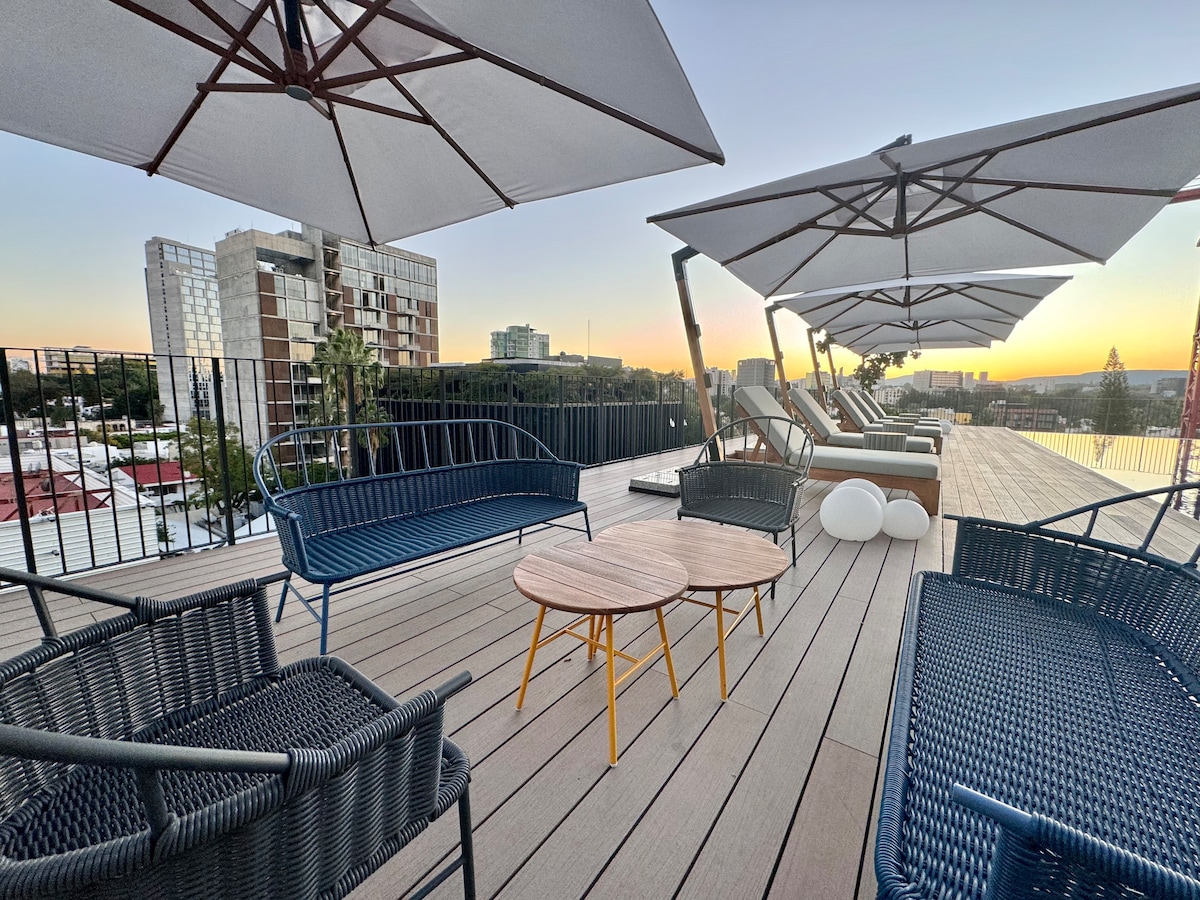 An outdoor terrace is featured with multiple loungers and shaded seating areas. Two wooden tables are positioned between the chairs, while large umbrellas provide shade. A city skyline is visible in the background, reflecting the colors of a sunset.