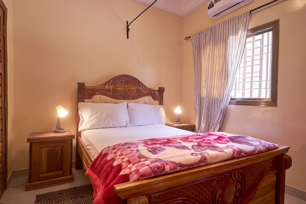 A comfortable bedroom showcases a wooden bed with soft bedding and decorative throw. Nightstands are positioned on either side, each with a lamp for ambient lighting. Natural light filters in through a window adorned with light curtains, adding a cozy touch to the space.