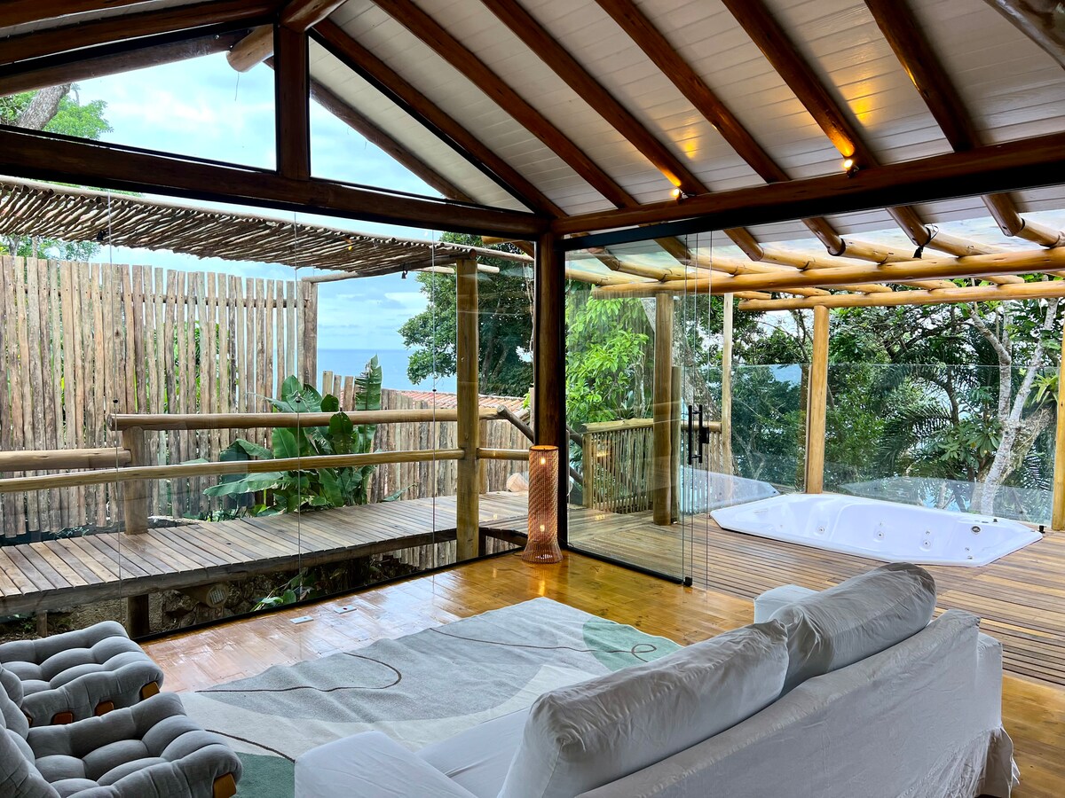 A spacious living area is framed by large glass walls, allowing natural light to fill the space. A comfortable sofa is positioned on a wooden floor, facing an outdoor terrace that provides views of the surrounding greenery and ocean. A hot tub is located nearby on the deck.