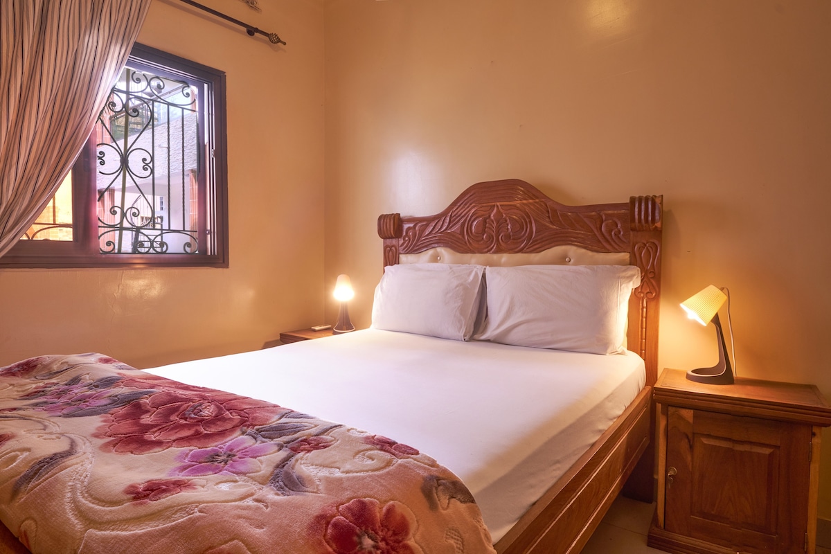 A cozy bedroom is featured, showcasing a spacious bed with floral bedding. Soft lighting from two bedside lamps creates a warm glow. The window is adorned with flowing curtains, and a decorative wrought iron grille is visible, offering a blend of privacy and light.