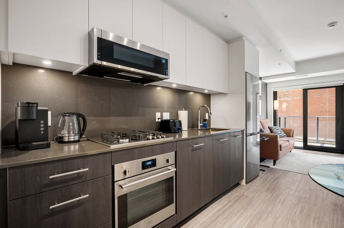 A modern kitchen is equipped with stainless steel appliances, including a gas stove and an oven, alongside a coffee maker and kettle. Sleek cabinetry complements the countertops. Natural light filters in through a nearby window, illuminating the open layout that seamlessly connects to the living area.