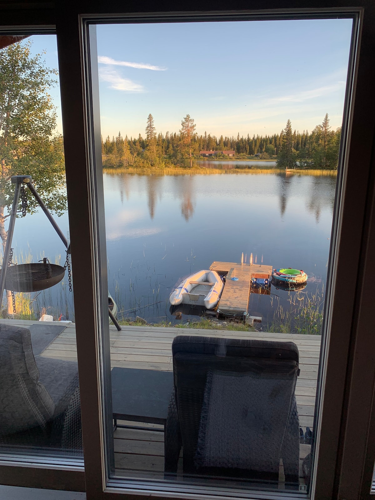 A serene view from a large glass door showcases the calm water reflecting the landscape. A wooden deck is adorned with two chairs, while a small dock features a rowboat and a kayak. Lush trees line the shore, creating a natural sense of tranquility.