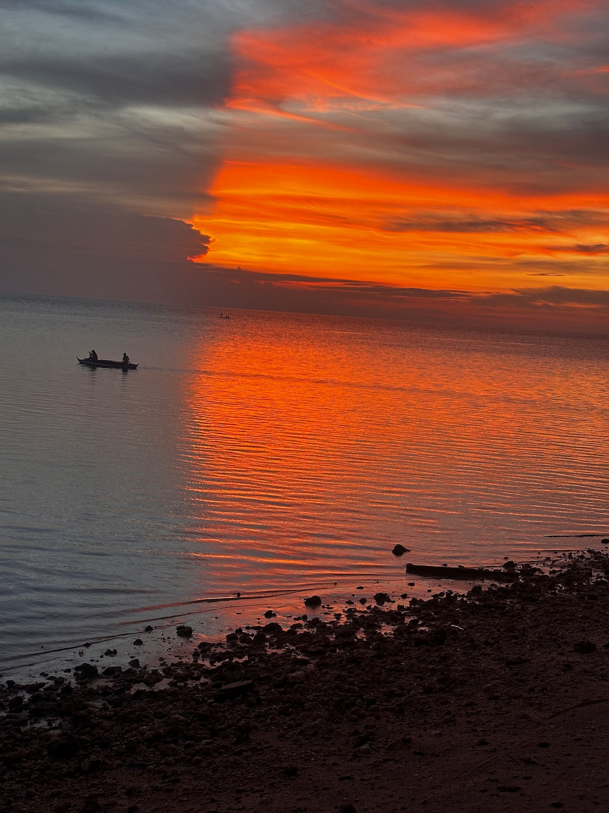 A vibrant sunset is captured over a calm body of water, reflecting rich orange and soft pink hues. Silhouetted against the colorful sky, a small boat is seen gently gliding across the water, creating a serene ambiance in the scene.