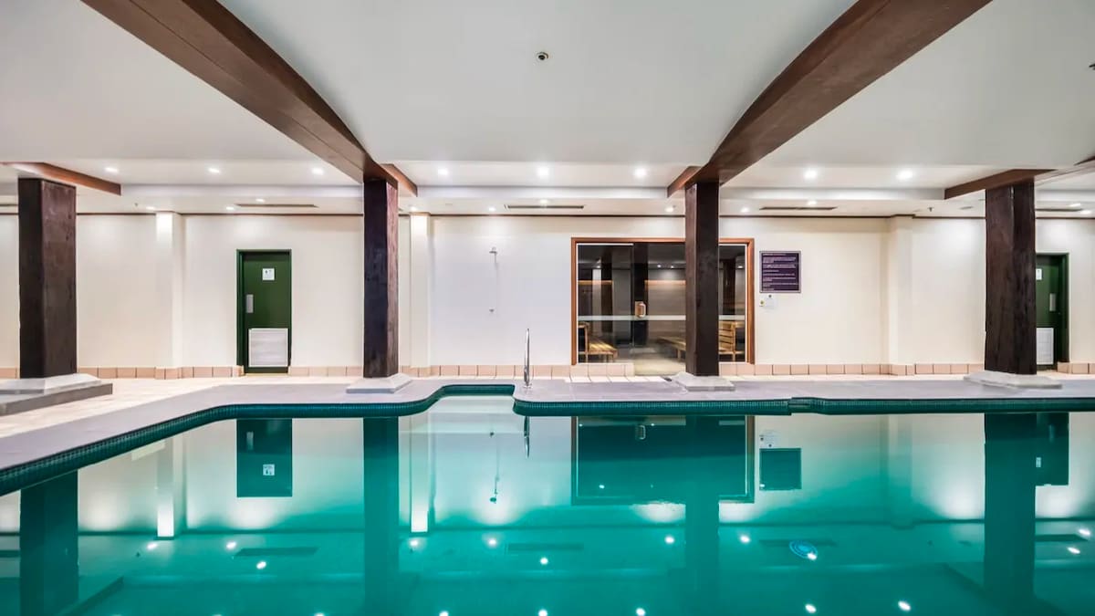 An indoor pool area showcases tranquil water reflecting bright lights above. The space is framed by elegant wooden beams and well-lit corners, alongside a seating area visible through expansive glass windows. The serene atmosphere invites relaxation and enjoyment.