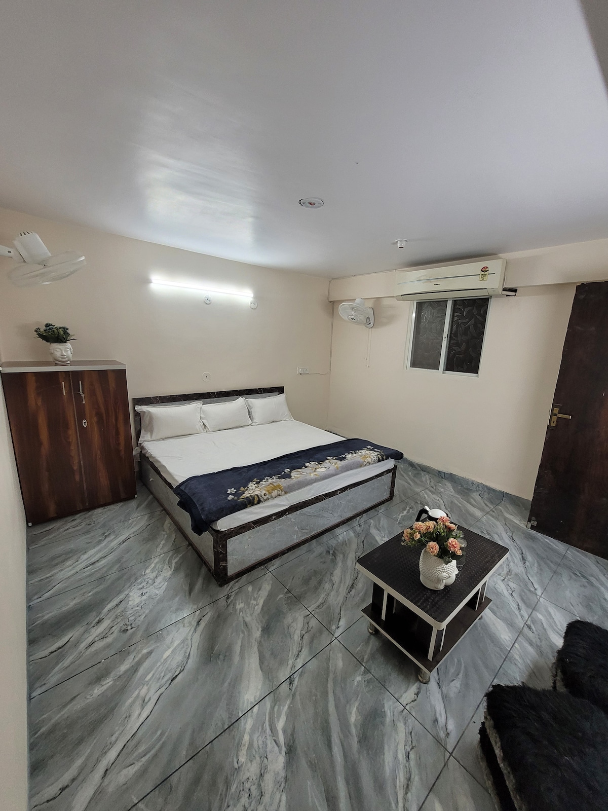 The room features a spacious bed adorned with decorative pillows and a floral arrangement on the cover. A small table with a vase of flowers occupies the center, while a window allows natural light to enter. Storage is provided by a wooden cabinet against the wall.