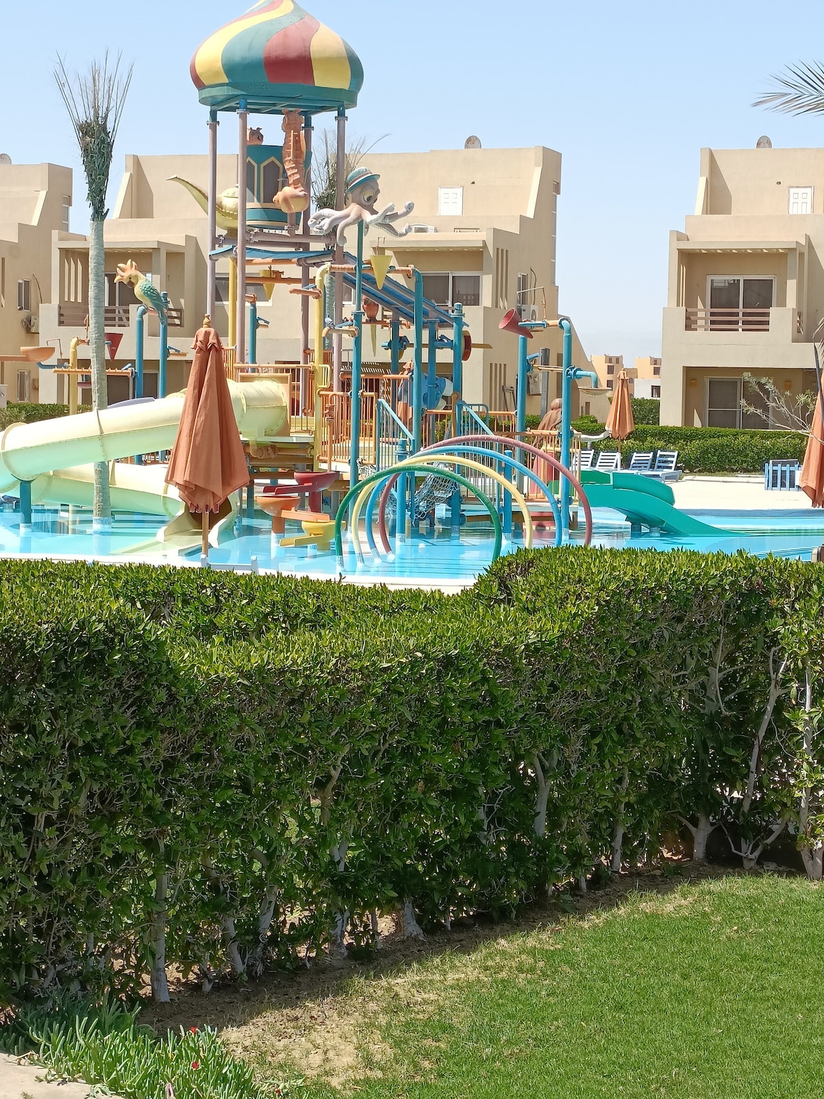 A vibrant water park area is featured, showcasing colorful slides and play structures surrounded by lush greenery. Sun loungers are positioned by the edge of the pool, and orange umbrellas provide shade for relaxation. The backdrop consists of chalets, adding to the inviting resort atmosphere.