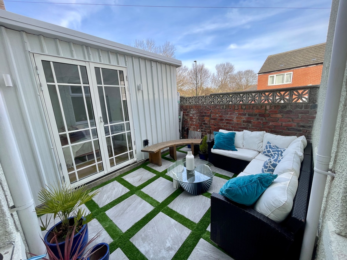 A small patio area features a comfortable outdoor seating arrangement, complemented by green cushions and a compact coffee table. Potted plants add a touch of nature to the space, while the surrounding wall of brick provides privacy and a cozy atmosphere.