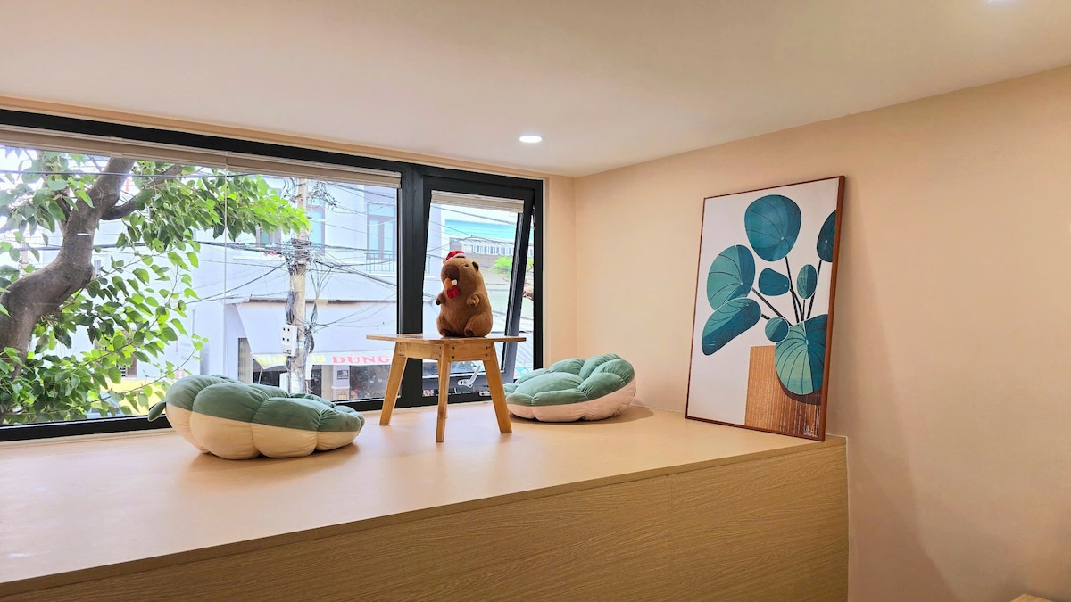 A bright and airy space features a wooden platform topped with a plush toy and a small table. Leafy art adorns the wall, complemented by two round cushions placed nearby. Large windows provide natural light, offering a view of greenery outside.