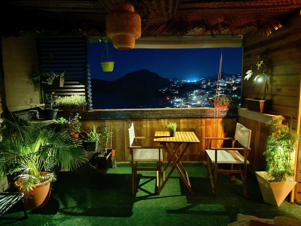 A cozy terrace is illuminated softly at night, featuring two wooden chairs and a small table surrounded by various potted plants. The view outside presents a cityscape with twinkling lights against a dark sky, creating a serene atmosphere.