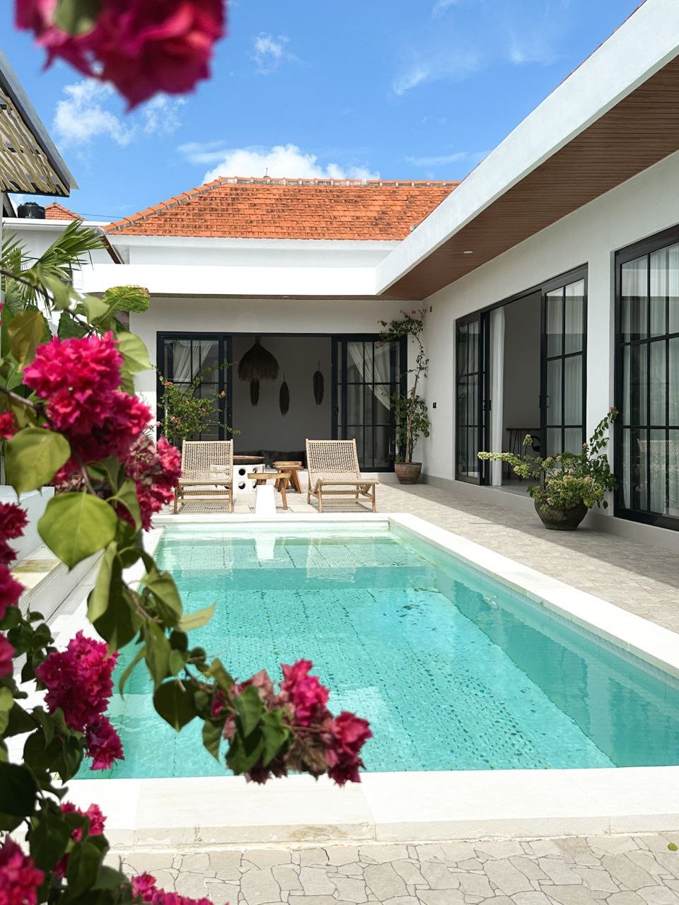 A serene private pool is framed by vibrant pink flowering plants, with two wooden lounge chairs placed nearby. Floor-to-ceiling glass doors offer a view into the bright living area, while the neutral tones of the surrounding architecture create a calming environment.