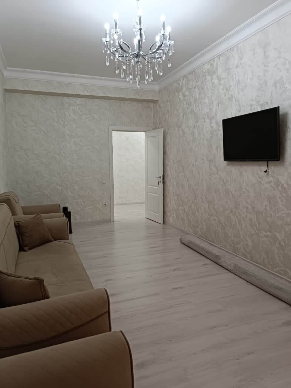 A spacious living area features a pair of comfortable beige sofas, light-colored wooden flooring, and a neutral textured wall. A flat-screen television is mounted on the wall, and a doorway leads to another room. A rolled carpet rests on the floor, adding to the decor.