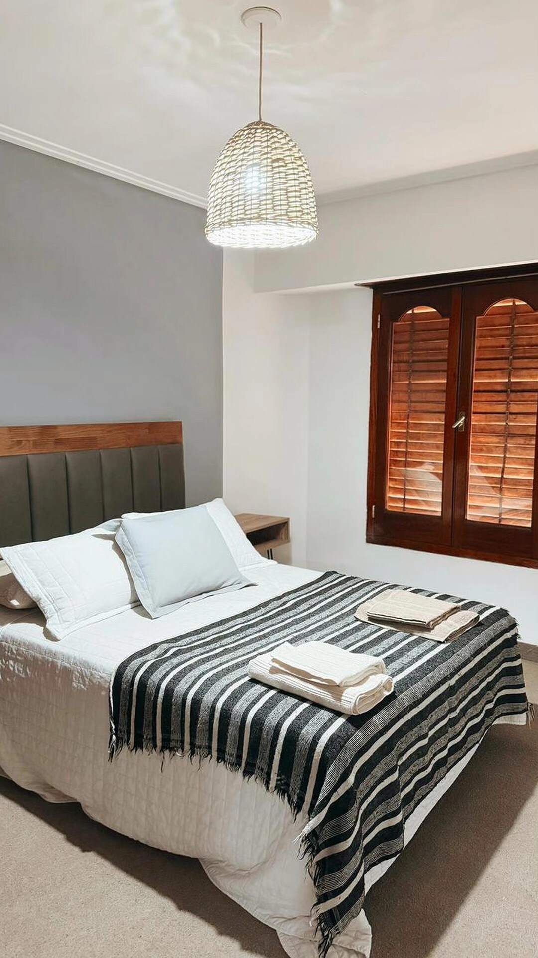 A bright bedroom features a spacious bed with a quilted white coverlet and decorative black and white striped throw. Soft natural light filters through wooden shutters, and a woven pendant light adds a touch of warmth to the space.