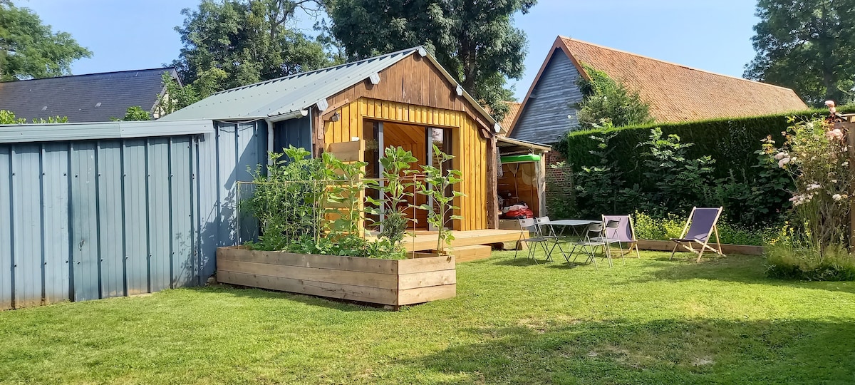 A small wooden cabin is surrounded by a lush garden featuring green plants and an outdoor seating area. A compact table with two chairs is visible beside the cabin. Nearby, a grey shed adds a contrasting element to the natural setting.