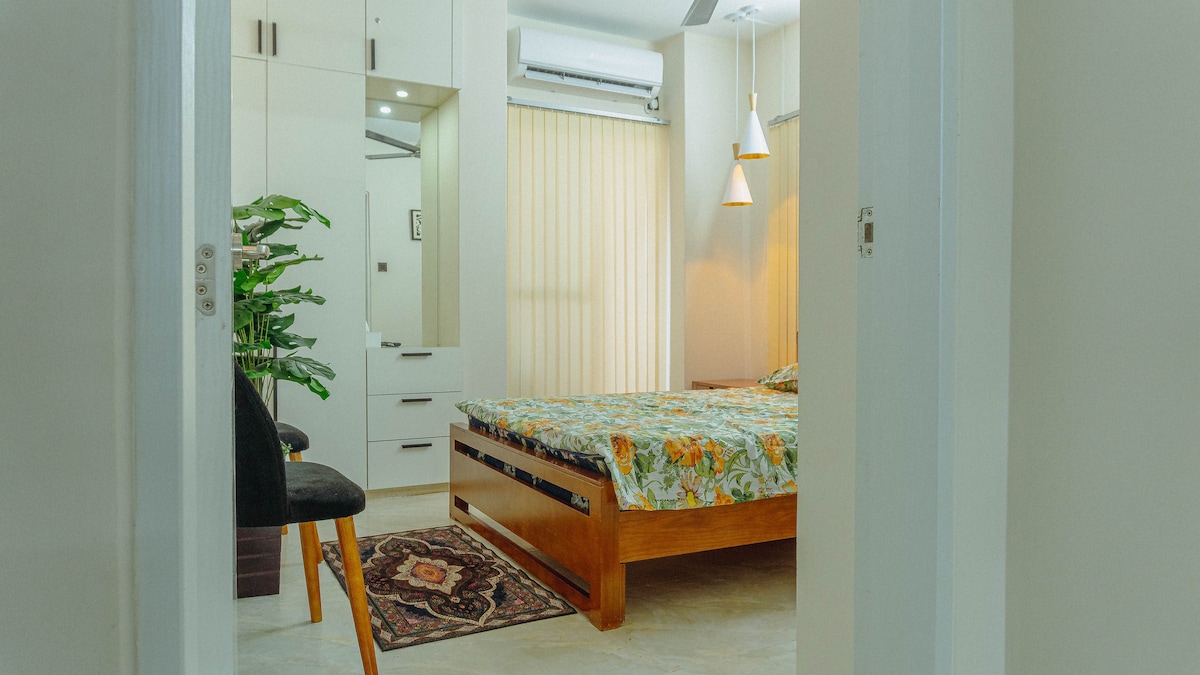 A well-lit sleeping area features a spacious bed topped with a floral patterned cover. Light yellow curtains allow soft light to filter in. A planter adds a touch of greenery, while stylish pendant lights hang above. A small area rug offers a cozy accent on the floor.
