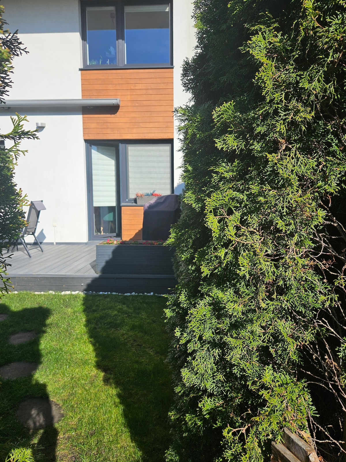 A well-maintained garden with lush greenery frames the view of a modern building. Beyond the garden, a wooden deck area is visible, showcasing comfortable seating and large windows that allow natural light to brighten the interior space.