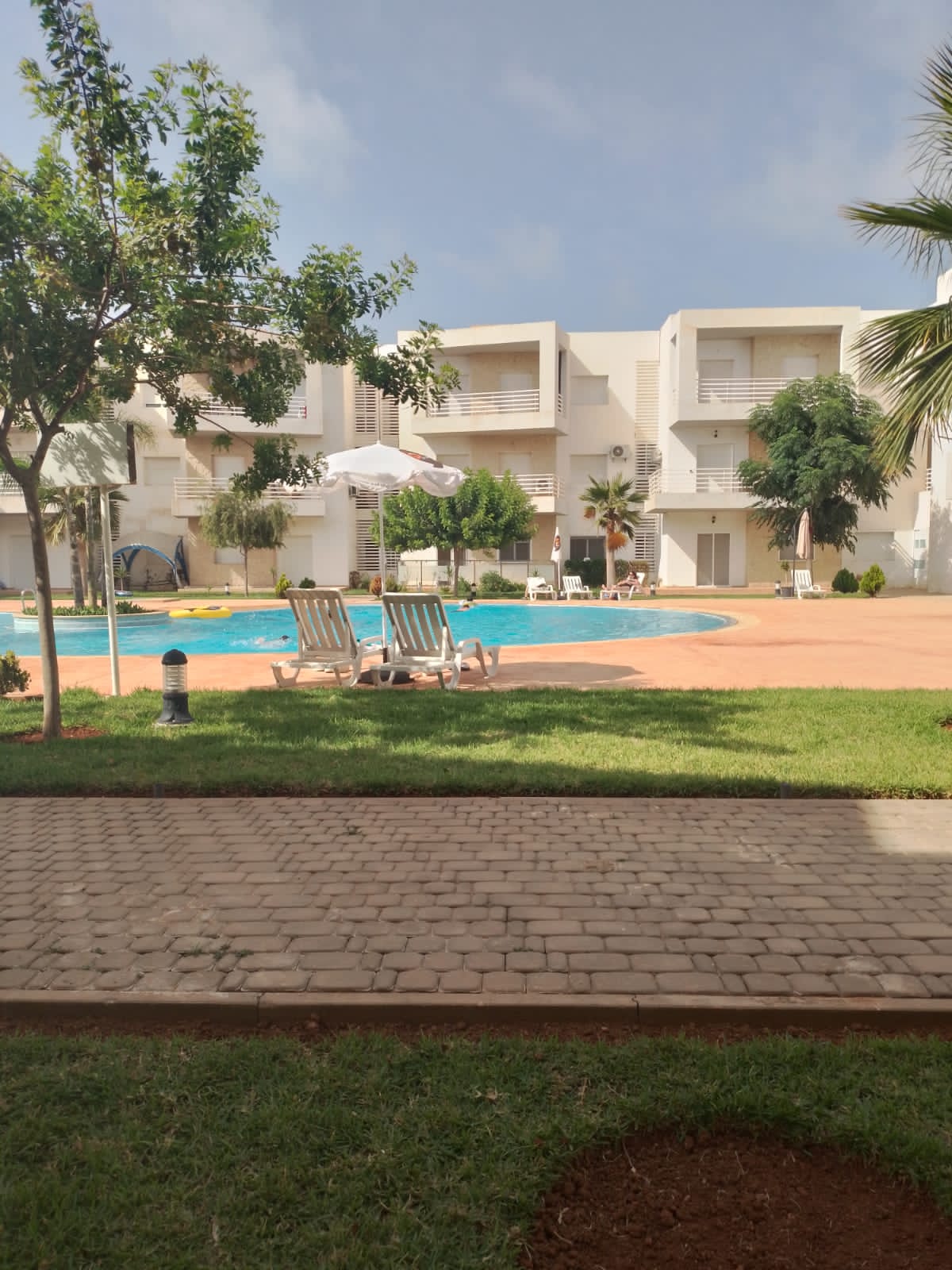 A serene outdoor area features a swimming pool surrounded by greenery. Sun loungers are positioned on a lawn, while a parasol provides shade. Nearby, palm trees add to the tropical atmosphere, with apartment buildings visible in the background.