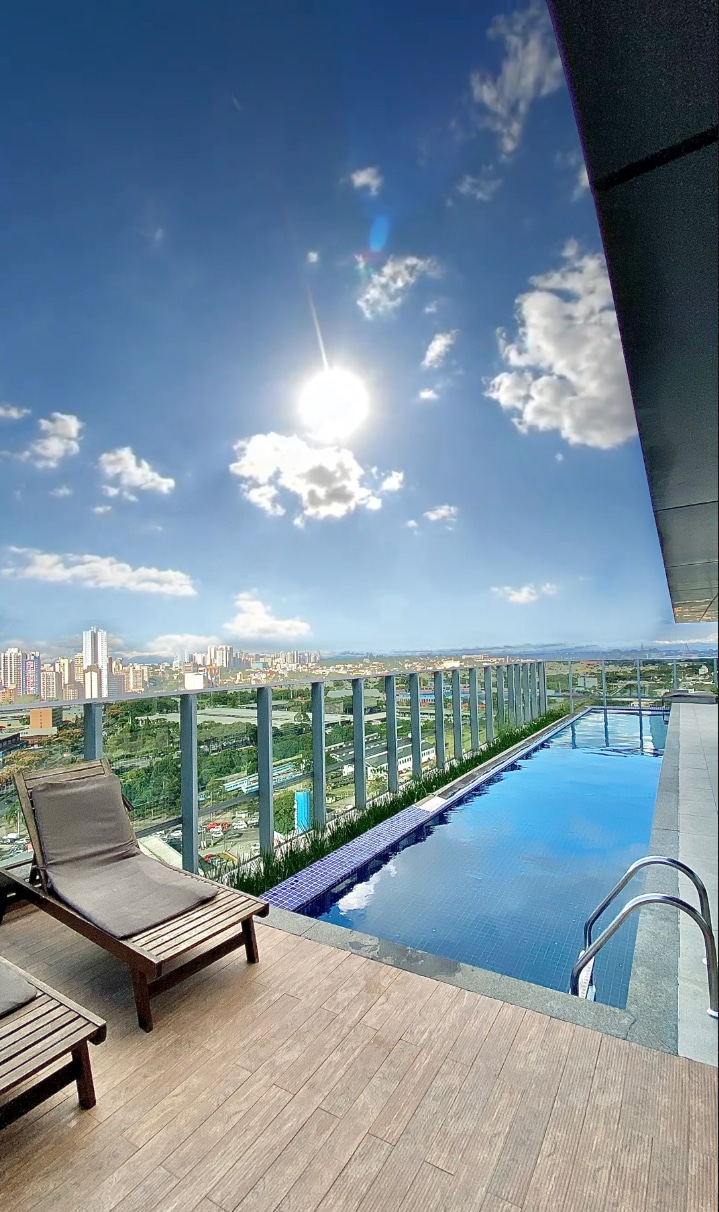 A rooftop area showcases a swimming pool with clear blue water reflecting the sky. Two lounge chairs are placed beside the pool, overlooking a panoramic city view. The sun shines brightly, casting a warm light on the scene and enhancing the vibrant atmosphere.