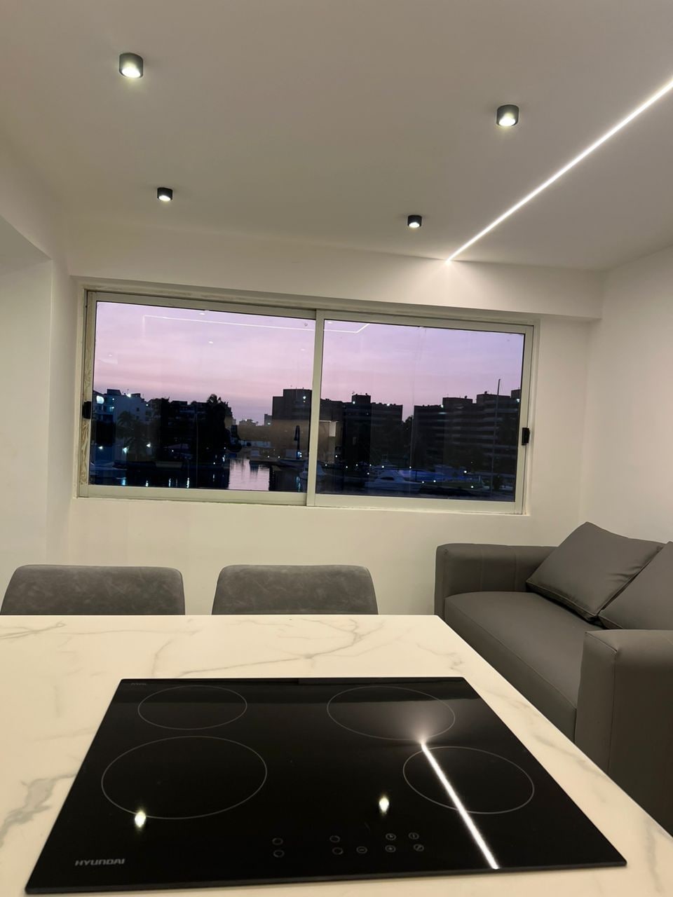 A modern kitchen space is visible, featuring a sleek induction cooktop atop a marble countertop. A cozy gray sofa is positioned nearby, with a large window offering a view of the city skyline at dusk, showcasing soft pink hues in the sky.