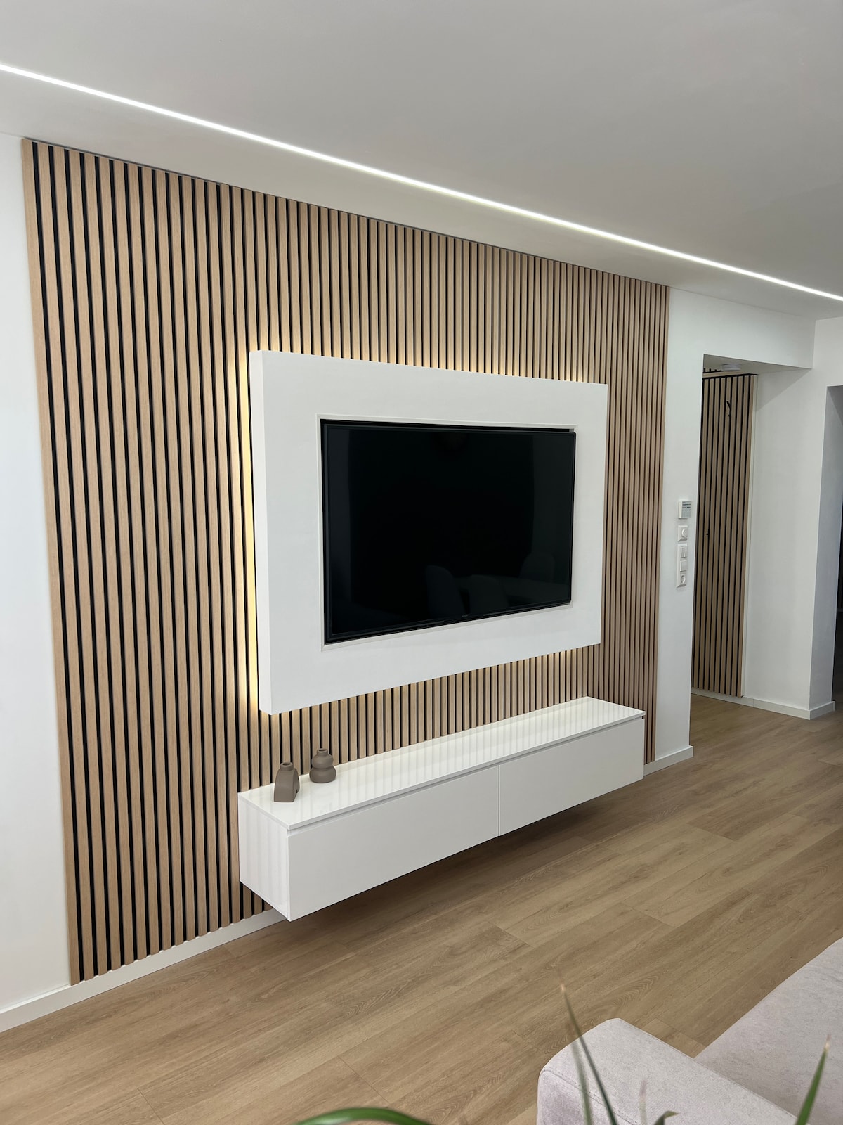 A modern living area features a wall-mounted television framed by a sleek white design. Below, a minimalistic white media console offers storage, complemented by decorative items. The wall is adorned with vertical wooden slats, adding texture to the overall aesthetic.