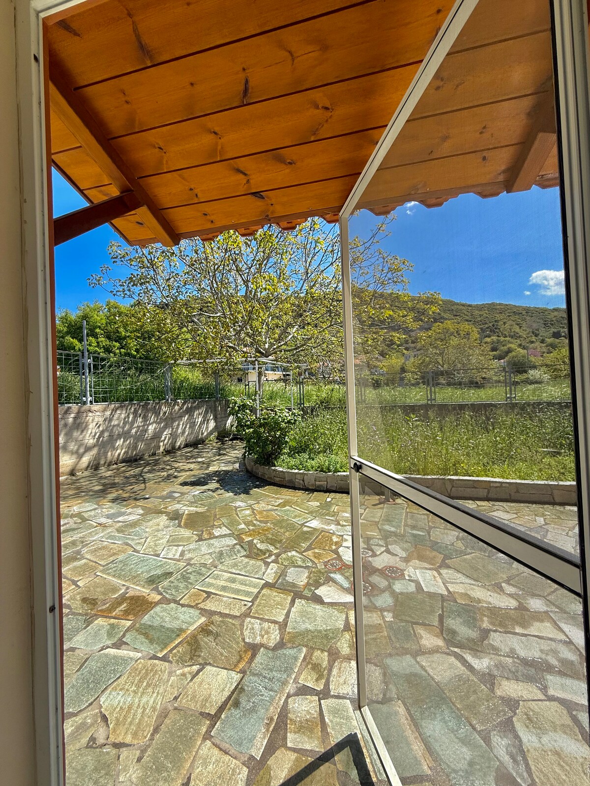A spacious, open doorway frames a serene outdoor area, highlighted by a textured stone patio. A lush green tree stands in the background, surrounded by gardens, with clear skies above, creating a bright and inviting ambiance for relaxation.